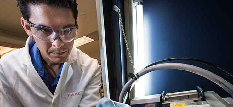 Photo of a man in a white lab coat, and protective glasses and gloves holding a glass microscope