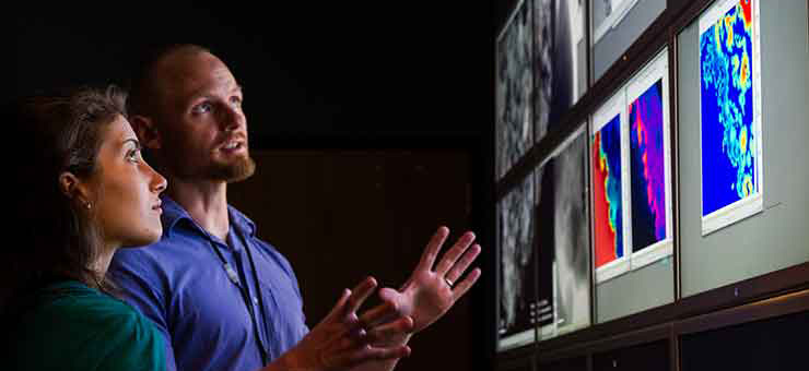 Photo of a woman and a man in a dark room looking at large monitor displays of images