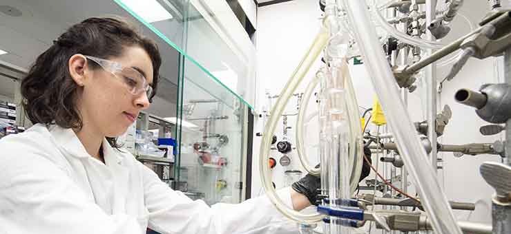 Photo of a woman in a white lab coat, and protective glasses and gloves using lab equipment