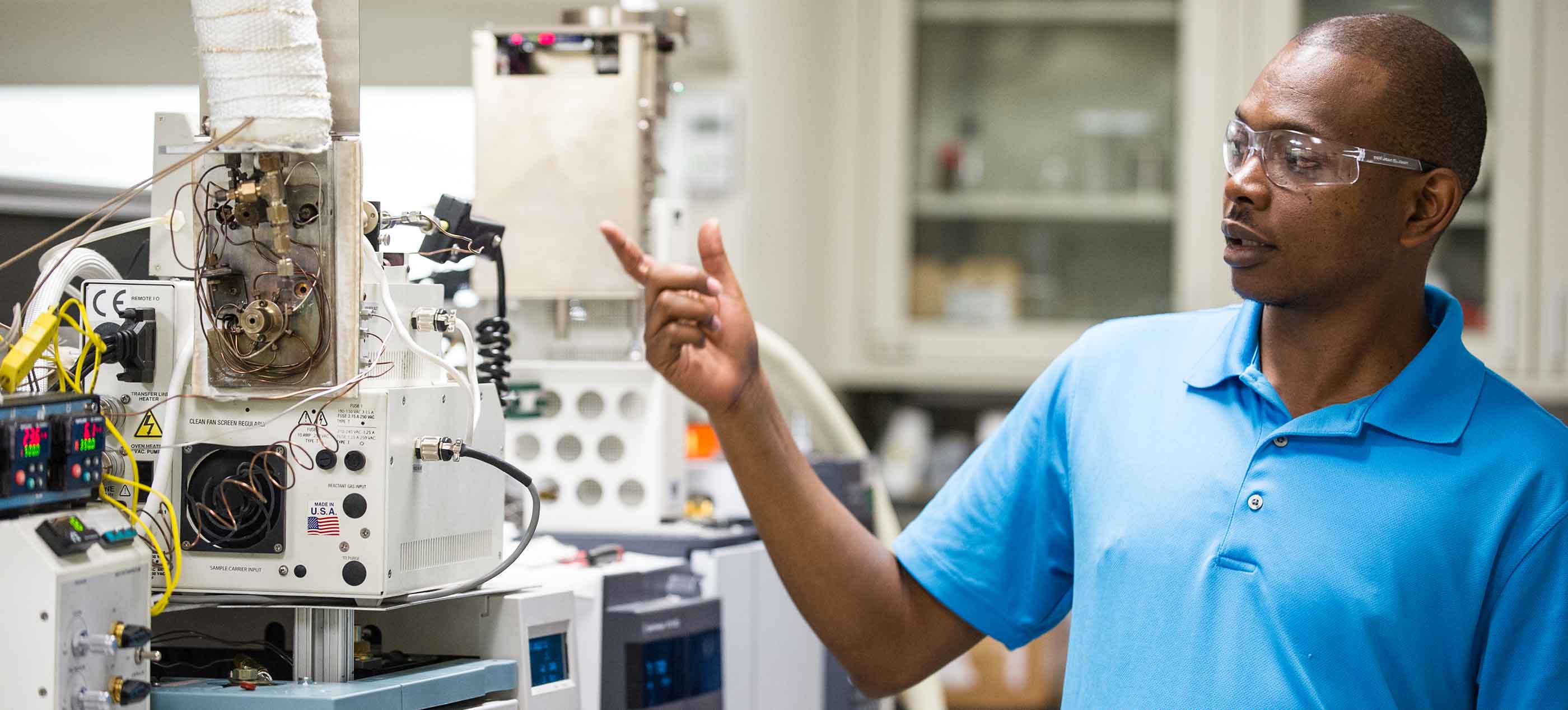 Photo of a woman in a white lab coat, and protective glasses and gloves using lab equipment