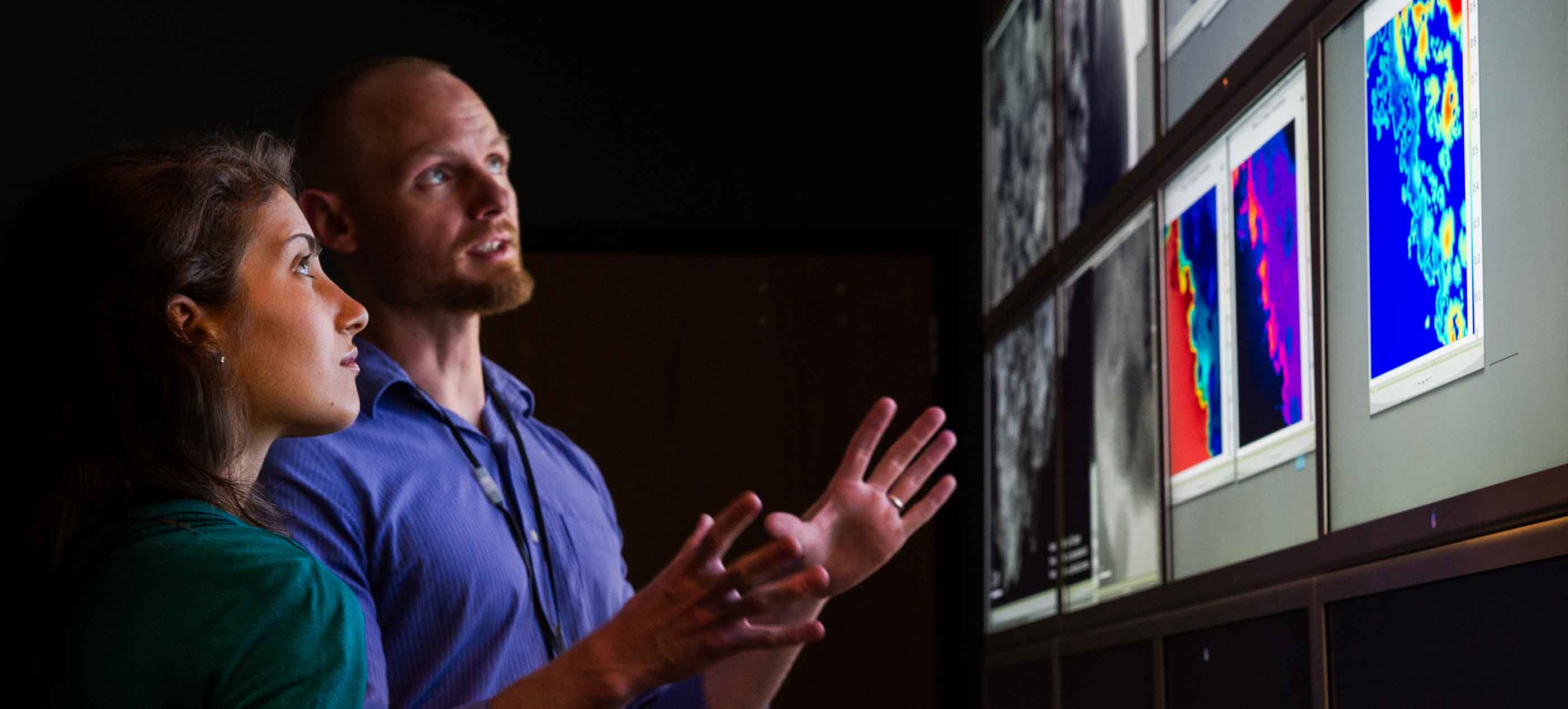 Photo of a woman and a man in a dark room looking at large monitor displays of images