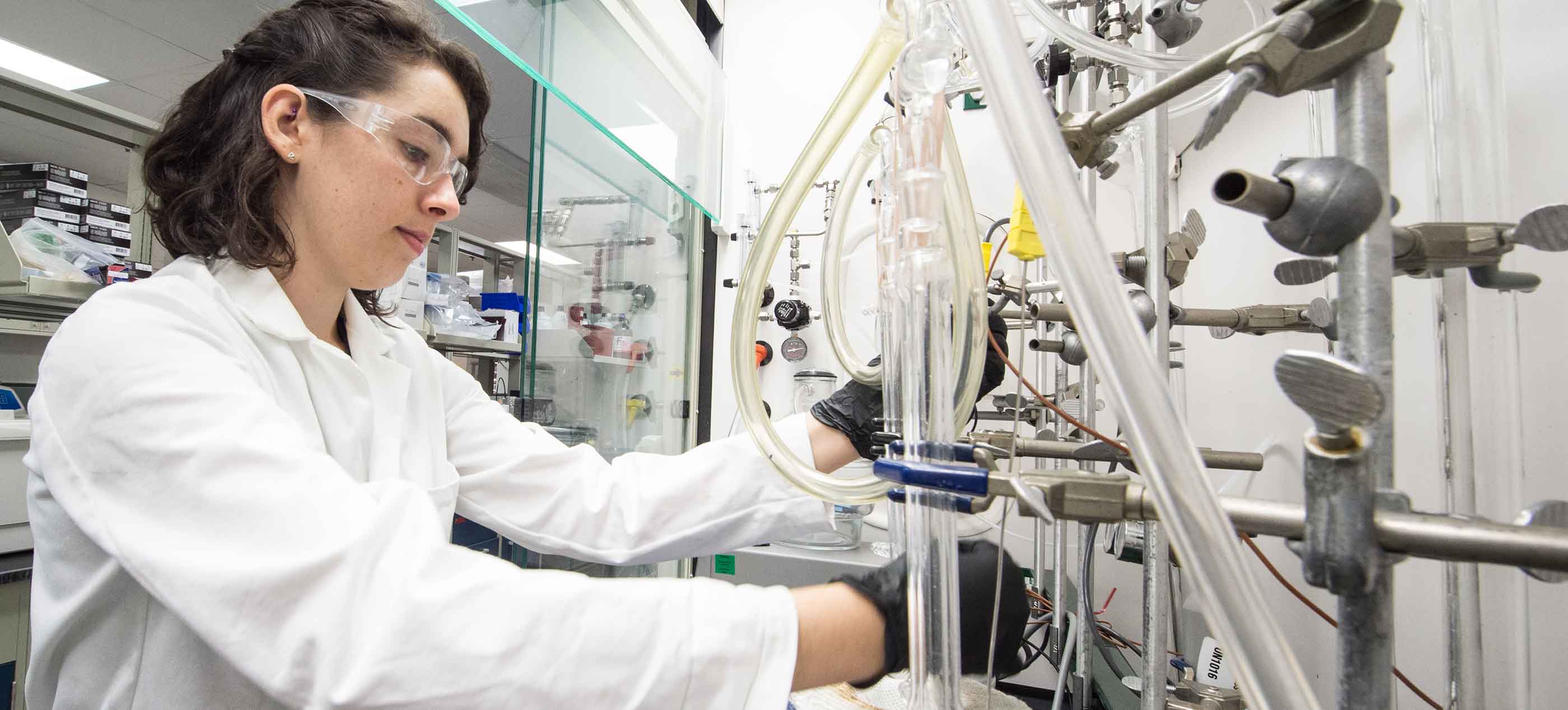 Photo of a woman in a white lab coat, and protective glasses and gloves using lab equipment