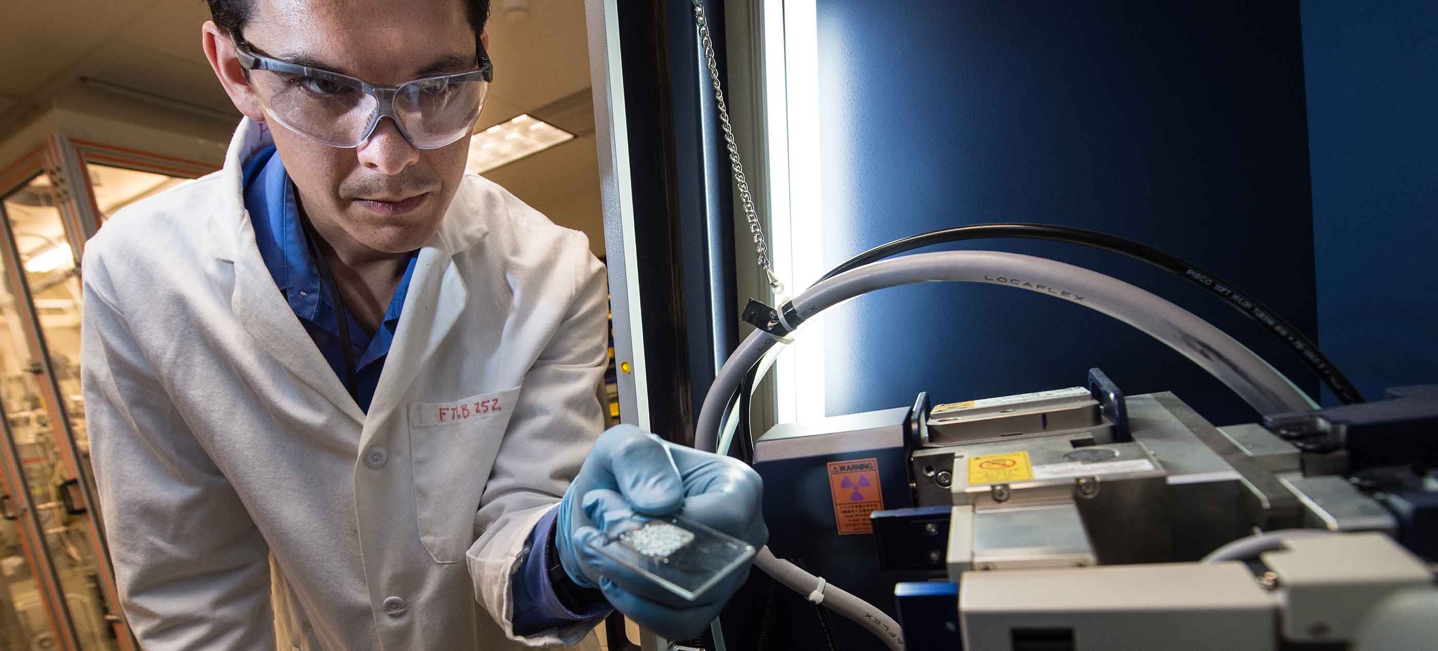 Photo of a man in a white lab coat, and protective glasses and gloves holding a glass microscope